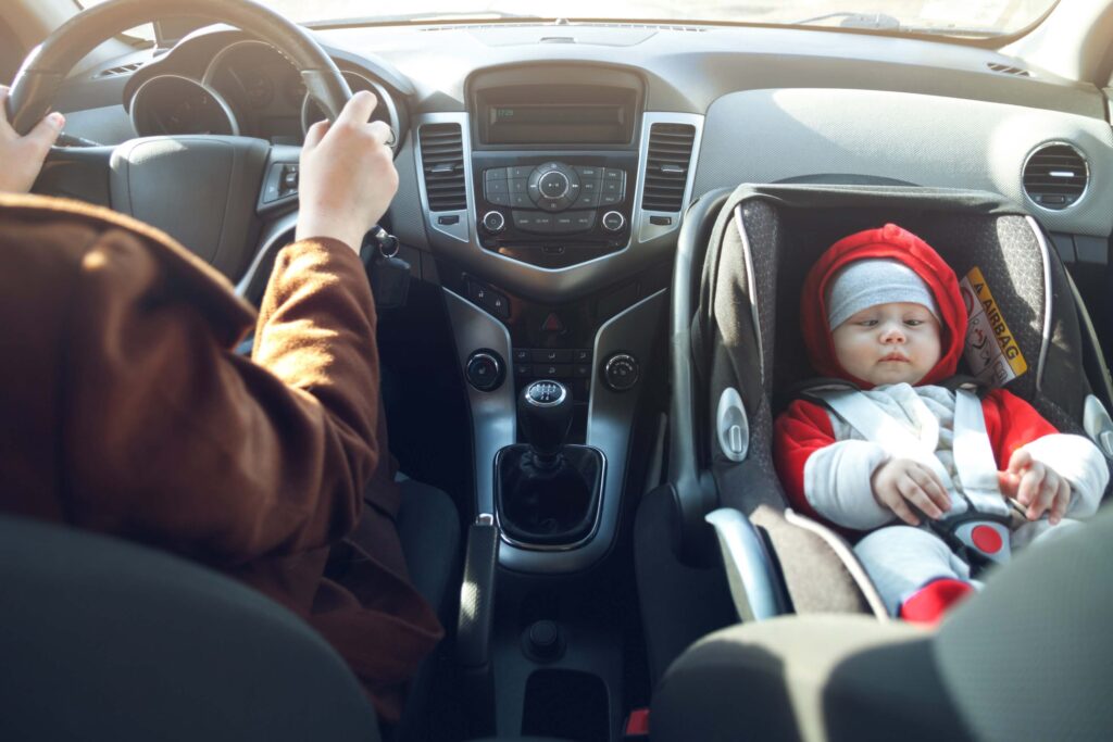 Woman drives the car, and her little child sits in the front baby car seat fastened by a seat belt.